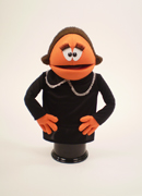 Orange puppet with brown hair and big eyes and large eyelashes.